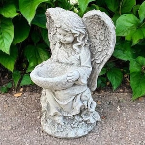 May include: A gray concrete angel statue with wings holding a bird bath. The angel is kneeling and has a sweet expression on its face.