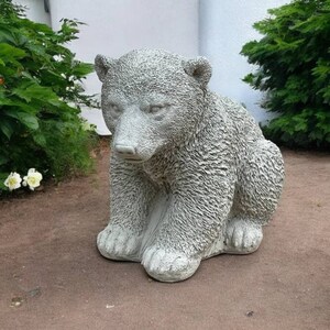 May include: A gray concrete sculpture of a bear cub sitting on the ground. The bear cub is detailed with fur and has a realistic appearance.