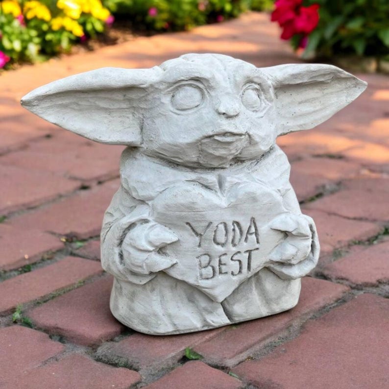 May include: A light gray concrete statue of a character holding a heart-shaped sign. The sign reads "YODA BEST". The statue has large ears and a detailed face. The statue is on a brick surface.