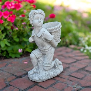 May include: A grey stone garden statue of a young child kneeling, carrying a woven basket on their back. The statue has detailed clothing and facial features. The statue is placed on a brick surface, with a background of green foliage and pink flowers.