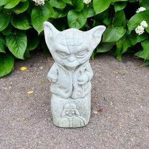 May include: A gray concrete statue of a character with large ears and a robe, resembling Yoda, with a Darth Vader figure at the base. The statue is set against a backdrop of green foliage and white flowers.