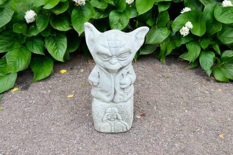 May include: A gray concrete statue of a character with large ears and a robe, resembling Yoda, with a Darth Vader figure at the base. The statue is set against a backdrop of green foliage and white flowers.