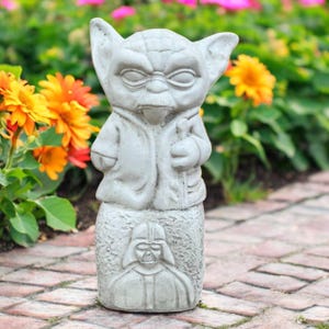 May include: A gray concrete garden statue of Yoda holding a staff, with Darth Vader's likeness on the base. The statue is surrounded by colorful flowers, including orange and yellow dahlias, and is set on a brick pathway.