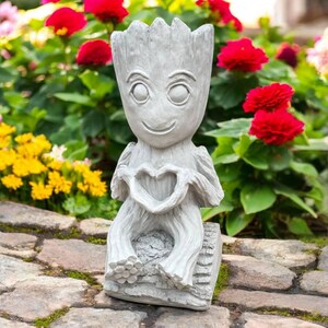 May include: A gray concrete garden statue of a smiling character holding a heart-shaped symbol. The statue has a tree-like appearance and is seated on a log-shaped base. The background features vibrant red and yellow flowers.