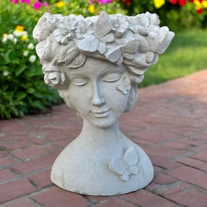 May include: A gray concrete planter shaped like a woman's head and shoulders. The woman has closed eyes and a serene expression. The planter is adorned with floral and butterfly details, including a butterfly on the shoulder. Ideal for garden decor.