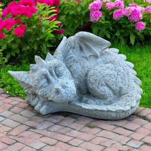 May include: A gray concrete dragon statue with wings, horns, and a textured body, resting on a brick surface. The dragon is surrounded by green grass and vibrant pink flowers, creating a whimsical garden ornament.