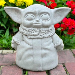 May include: A gray concrete statue of a character with large ears and a hooded coat. The statue has large eyes and a neutral expression. The statue is outdoors, surrounded by red, yellow, and green plants.
