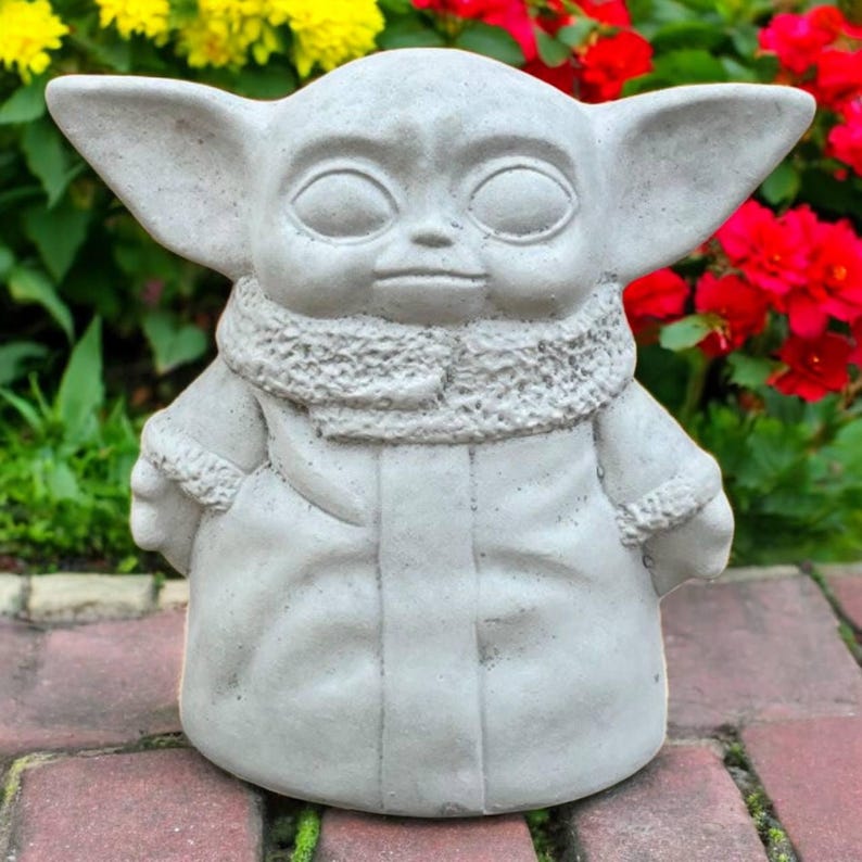 May include: A gray concrete statue of a character with large ears and a hooded coat. The statue has large eyes and a neutral expression. The statue is outdoors, surrounded by red, yellow, and green plants.