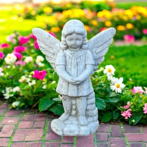 May include: A gray stone angel statue with outstretched wings, standing on a brick path. The angel is wearing a tunic and has its hands clasped in front. The background features colorful flowers and green grass.