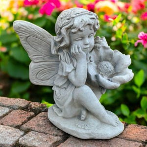 May include: A gray concrete fairy statue with detailed wings, a floral crown, and a serene expression. The fairy is seated, holding a large flower, and is suitable for garden decor.