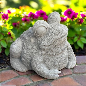 May include: A detailed, gray concrete frog statue with a textured surface. The frog is sitting on a brick surface, with a backdrop of vibrant pink flowers and green foliage. The statue has large, round eyes and a detailed skin texture.