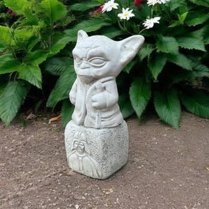 May include: A light gray Yoda statue with large ears and a robe, standing on a square base with a Darth Vader image. The statue is outdoors, surrounded by green plants and flowers, and is made of stone.