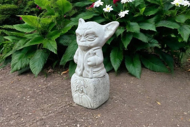 May include: A light gray Yoda statue with large ears and a robe, standing on a square base with a Darth Vader image. The statue is outdoors, surrounded by green plants and flowers, and is made of stone.