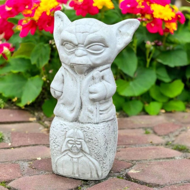 May include: A gray concrete Yoda statue with a Darth Vader design on the base. The statue is in front of a backdrop of red, yellow, and green flowers. The statue is a decorative garden ornament.