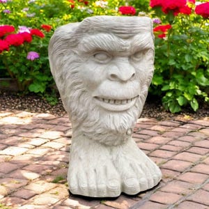 May include: A light gray Bigfoot planter with a smiling face and a large foot base. The planter is made of a stone-like material and is designed to hold plants. The background features a garden with red and pink flowers.