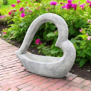 May include: A gray concrete heart-shaped planter with a cutout in the center. The planter is sitting on a brick walkway in a garden setting.