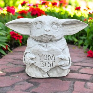 May include: A light gray statue of a character with large ears, holding a heart-shaped sign that reads "YODA BEST". The statue is outdoors, set against a backdrop of red flowers and greenery.