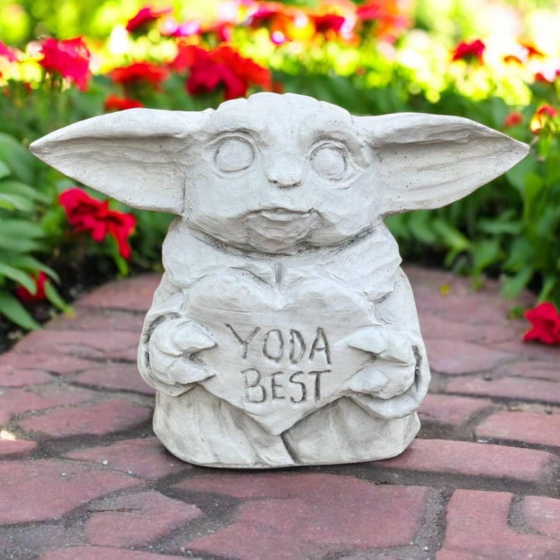 May include: A light gray statue of a character with large ears, holding a heart-shaped sign that reads "YODA BEST". The statue is outdoors, set against a backdrop of red flowers and greenery.