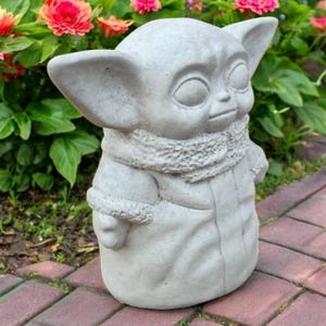 May include: A gray concrete statue of a character with large ears and a scarf. The statue is outdoors, surrounded by green foliage and red and orange flowers. The statue is a decorative garden ornament.