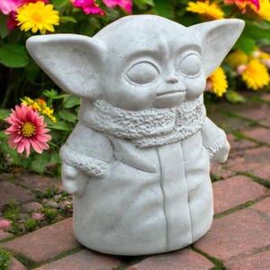 May include: A light gray concrete statue of a character with large ears and a textured scarf. The statue is standing on a brick surface, surrounded by colorful flowers in shades of yellow, pink, and green. The statue is a popular character from a science fiction franchise.