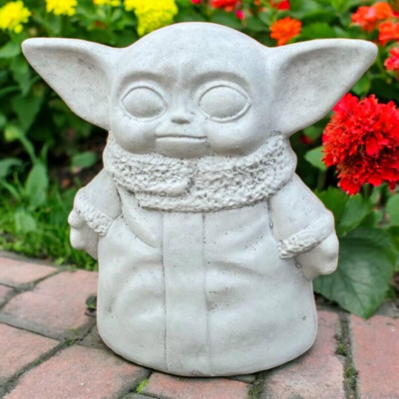 May include: A light gray concrete statue of a character with large ears, large eyes, and a textured scarf. The statue is wearing a coat with a textured collar and cuffs. The statue is outdoors, surrounded by red, yellow, and green plants.