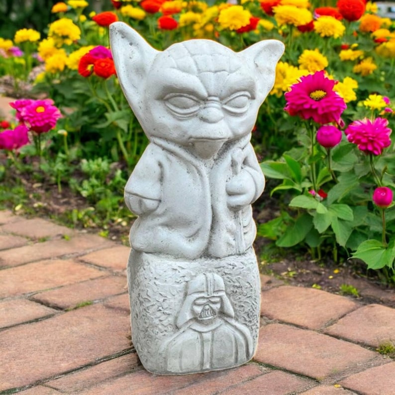May include: A gray concrete garden statue featuring Yoda on top and Darth Vader below. The statue is set against a backdrop of colorful flowers, including pink, yellow, and red blooms, and a brick pathway.
