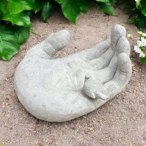 以下が含まれることがあります： 手のひらに鳥がとまっているコンクリート製のハンド彫刻。手は砂利の上に置かれています。