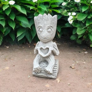 May include: A gray concrete garden statue of a character from the movie Guardians of the Galaxy. The character is sitting with its hands forming a heart shape.