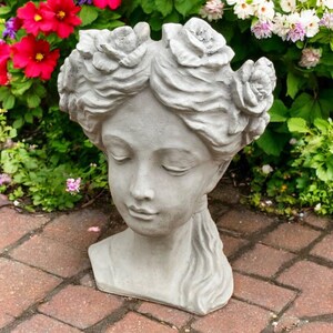 May include: A gray concrete garden statue of a woman's head, adorned with rose-shaped flowers in her hair. The statue has closed eyes and a serene expression. The head is set on a brick patio, with colorful flowers and greenery in the background.
