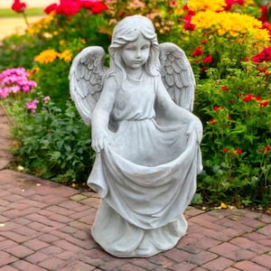 Peut inclure: Statue d'ange grise avec des ailes déployées et une expression sereine. L'ange tient les bords de sa robe, créant une forme de bassin. La statue est posée sur un patio en briques, entourée de fleurs colorées.