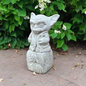 May include: A gray concrete statue of a character with large ears and a robe, standing on a block with a Darth Vader image. The statue is outdoors, set against a backdrop of green foliage and white flowers.
