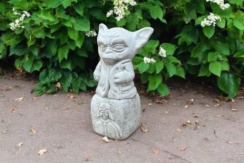 May include: A gray concrete statue of a character with large ears and a robe, standing on a block with a Darth Vader image. The statue is outdoors, set against a backdrop of green foliage and white flowers.