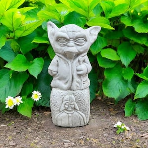 May include: A gray concrete statue of Yoda from Star Wars, standing on a base with Darth Vader's likeness. The statue is surrounded by green foliage and small white and yellow flowers.
