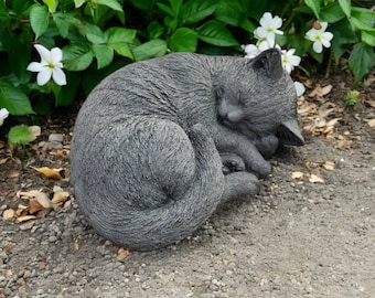 May include: A gray concrete sculpture of a cat curled up sleeping. The sculpture is placed on a bed of gravel and surrounded by green plants and white flowers.