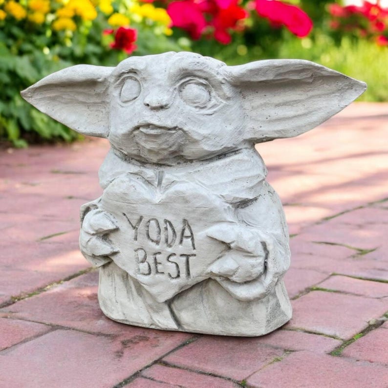 May include: A gray concrete statue of a character holding a heart-shaped sign that reads "YODA BEST." The statue has large ears and a serene expression. The statue is on a brick surface with flowers in the background.