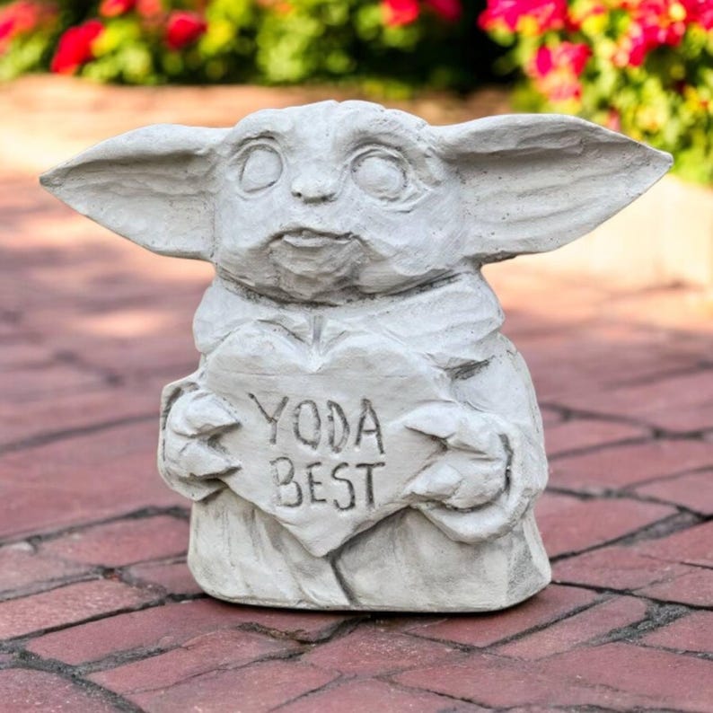 May include: A light gray statue of a character holding a heart-shaped sign. The sign reads "YODA BEST". The statue has large ears and a detailed face. The statue is sitting on a brick surface.
