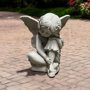 May include: A gray stone sculpture of a fairy with wings sitting with her head down. The fairy is wearing a dress and has flowers in her hair.