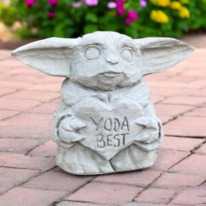 May include: A light gray statue of a character with large ears, holding a heart-shaped sign. The sign reads "YODA BEST". The statue is made of a stone-like material and is placed on a brick surface.