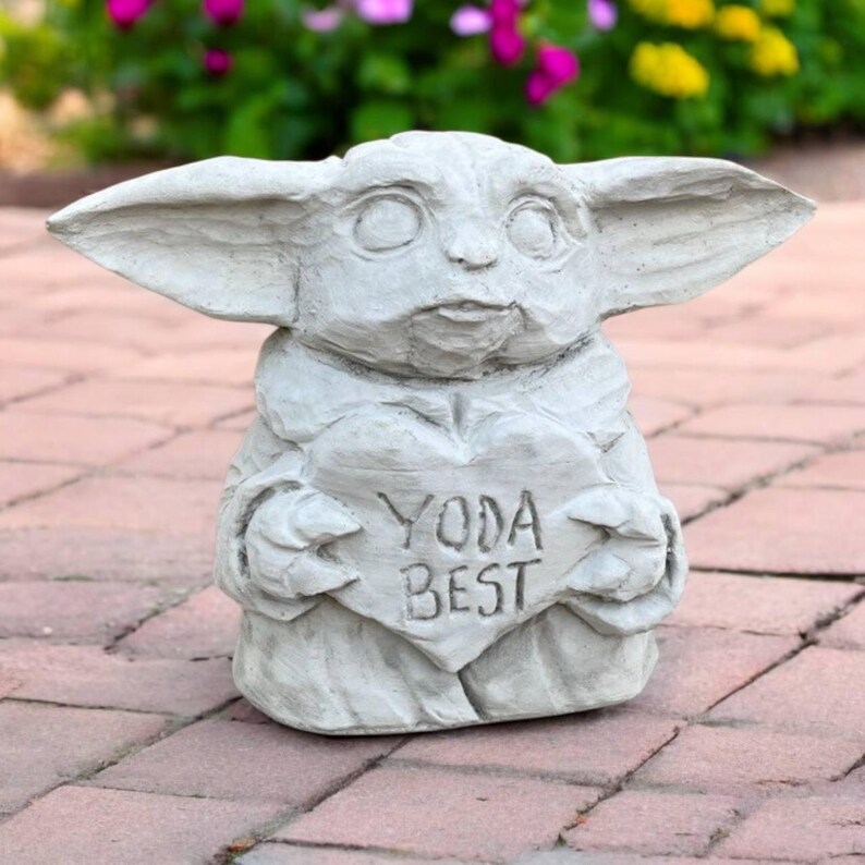 May include: A light gray statue of a character with large ears, holding a heart-shaped sign. The sign reads "YODA BEST". The statue is made of a stone-like material and is placed on a brick surface.