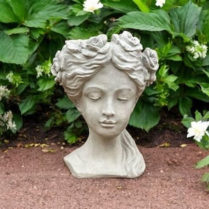 May include: A grey concrete planter in the form of a woman's head, decorated with rose-like flowers around the crown. The sculpture features closed eyes and a calm expression. The planter is set against a backdrop of green foliage and white flowers.