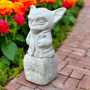 May include: A gray concrete Yoda statue with a detailed face and robe, holding a staff. The statue sits atop a base with a carved image of Darth Vader. The statue is surrounded by colorful flowers.