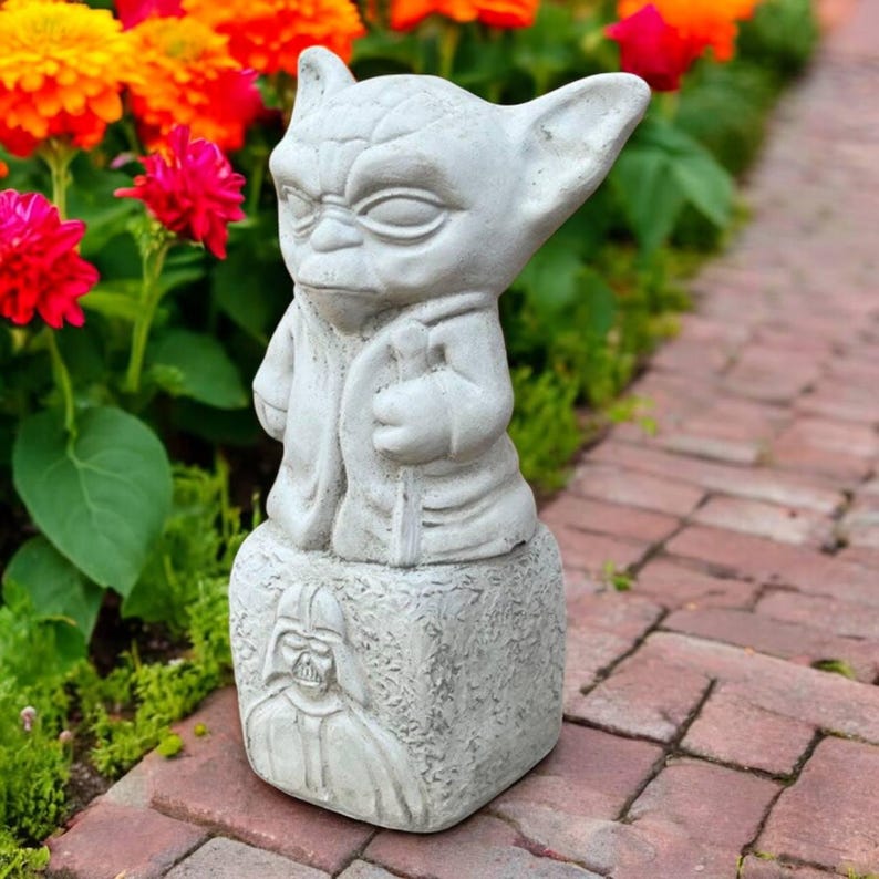 May include: A gray concrete Yoda statue with a detailed face and robe, holding a staff. The statue sits atop a base with a carved image of Darth Vader. The statue is surrounded by colorful flowers.