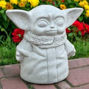 May include: A light gray concrete statue of a character with large ears, large eyes, and a textured scarf. The statue is standing on a brick surface with a background of red and yellow flowers and green foliage. The statue is a garden ornament.