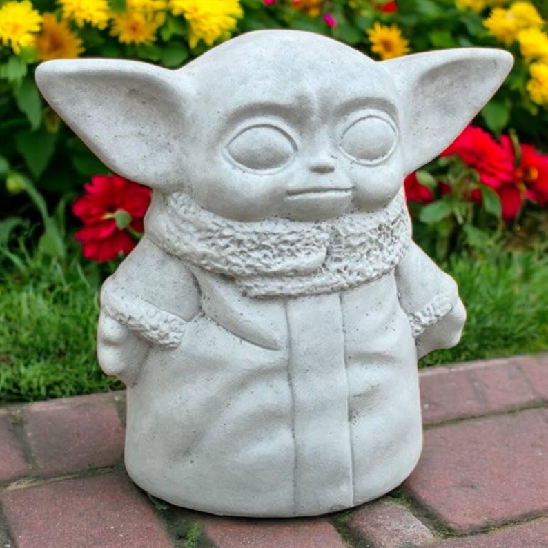 May include: A light gray concrete statue of a character with large ears, large eyes, and a textured scarf. The statue is standing on a brick surface with a background of red and yellow flowers and green foliage. The statue is a garden ornament.