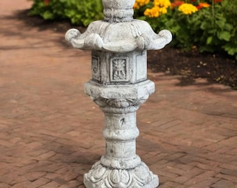 May include: A gray stone Japanese garden lantern with a pagoda-style top and ornate carvings. The lantern stands on a hexagonal base and is surrounded by a brick walkway and colorful flowers.