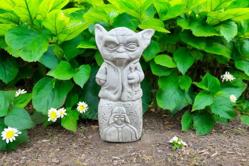May include: A gray concrete garden statue featuring Yoda on top and Darth Vader below. The statue is surrounded by green foliage and small white and yellow flowers.