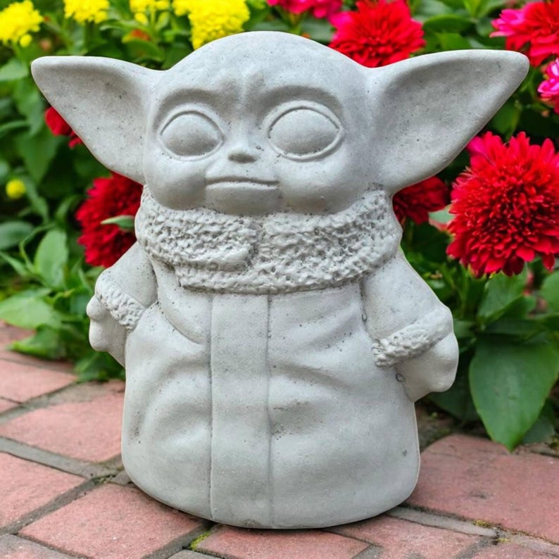 May include: A gray concrete statue of a character with large ears and large eyes, wearing a coat with a fur-like collar. The statue is outdoors, surrounded by red, yellow, and pink flowers.