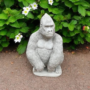 May include: A gray concrete gorilla statue sitting on a gravel surface. The gorilla is in a seated position with its arms resting on its legs.