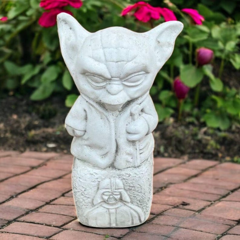May include: A light gray Yoda statue with a carved Darth Vader base. The Yoda figure is holding a staff and wearing a robe. The statue is made of a stone-like material and is suitable for outdoor display.