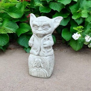 May include: A light gray Yoda statue with a detailed face and robe, standing on a base with a Darth Vader image. The statue is outdoors, surrounded by green foliage and white flowers, creating a garden decor theme.
